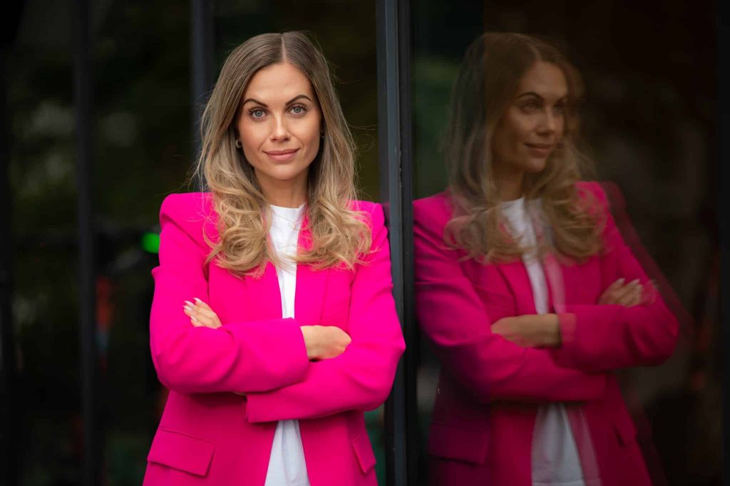 Dame med blondt hår og rosa blazer lener seg mot en glassvegg og ser inn i kamera